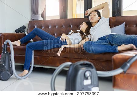 Asian couple tired to cleaning floor with vaccuum cleaner sleep on a brown sofa at home.