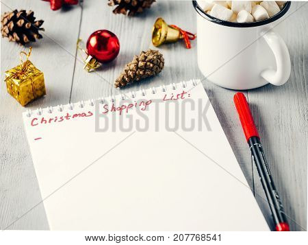Christmas gifts shopping planning. Make shopping or to-do list for Christmas. Notebook, mug hot chocolate with marshmallows, New Year's decoration and pine cone on gray wooden background.