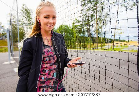 Smiling sporty woman listening music while holding mobile phone by fence
