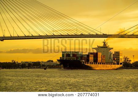 Container cargo ocean ship float on river and pass under bridge at sunset twilight with red sky