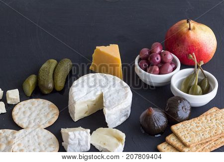 Cheese crackers pear dill pickles olives figs and caper berries on black background with space for text