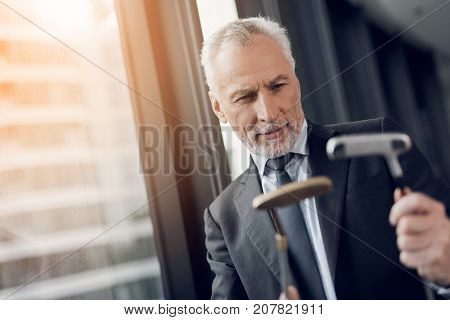 A respectable elderly man playing a mini golf in the office. He considers two golf clubs and wants to choose one of them for the upcoming game