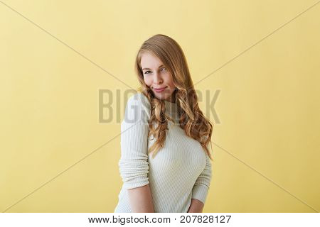 Isolated portrait of gorgeous stunning Caucasian female model with long curly hair looking at camera with playful flirty smile while posing for pciture during photo shooting at yellow studio wall