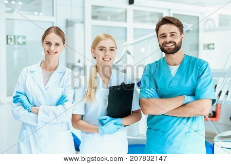 A dentist and two nurses are posing in the dental office on the background of dental equipment.