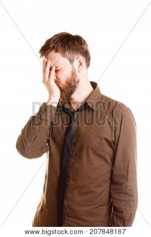 Shy young brunette man with beard in brown shirt and black tie covers face with palm on camera in studio isolated on a white background.