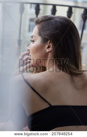 Vertical tender portrait of beautiful woman posing indoors through white curtain looking outside from indoor. Young brunette lady touching her softest skin feeling the moment enjoying fresh morning.