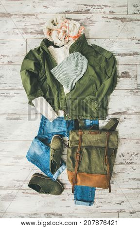 Flat-lay of Fall lady' s clothes outfit. Ecru colored knitted woolen sweater, parka, linen scarf, denim jeans, haki suede boots, backpack, cap over parquet background, top view. Trendy Autumn look