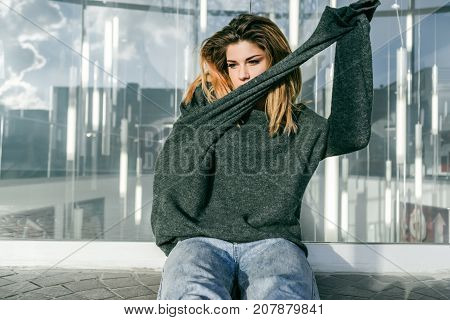 a girl in a big gray sweater takes it off and sits on a city street