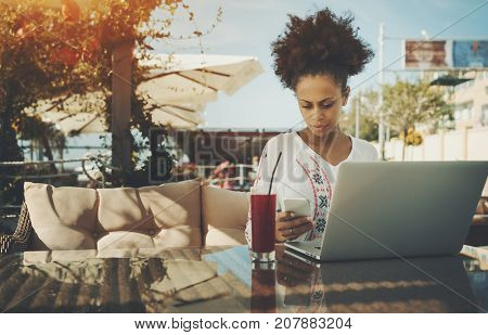 Serious young black female freelancer answering incoming phone message while sitting in street bar or cafe and working on project using laptop with copy space place for logo your text or advertising