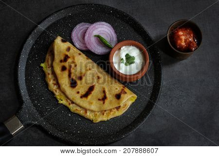 Aloo Paratha - Indian potato stuffed wheat bread served with Raita and mango pickle on dark moody background Top down view