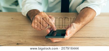 Smartphone in hands of successful businessman. Adult caucasian business person in white shirt with tucked rolled up sleeves using mobile phone at the desk in office.