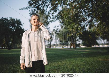 Young and good-looking student is standing in a beautiful green park this summer evening. He desided to have a walk after the lessons at university. Now this man is talking on the phone with somebody, smiling and enjoying the conversation.