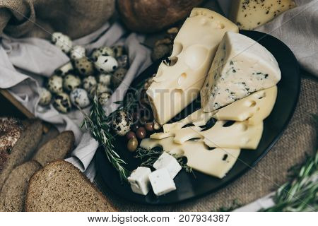 Different kind of cheese - blue cheese, cheese with holes, cheese slices and cubes on black plate. Slices and whole bread on beige background. Herbs and berries as decoration