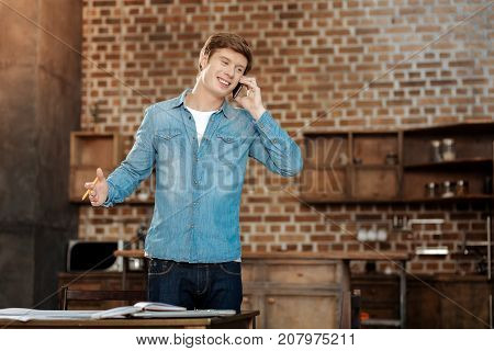 Great news. Cheerful young man standing near the table and talking on the phone while receiving pleasant news from him
