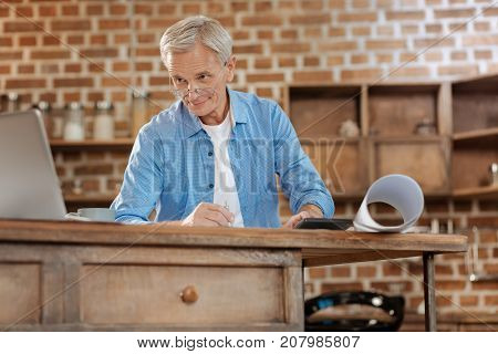 New information. Grey-haired senior engineer in spectacles standing near a kitchen counter, holding a compass and looking at the laptop screen