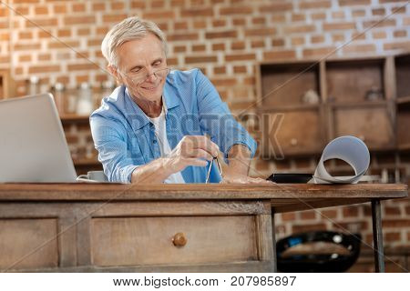 Precise data. Pleasant cheerful senior engineer in spectacles standing near a kitchen counter and measuring a distance on the blueprint with a pair of compasses