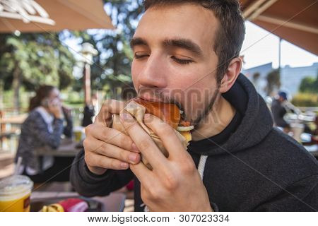 A Guy Is Greedily Eating His Burger While Sitting At The Restauraunt Terrace. His Eyes Are Closed.
