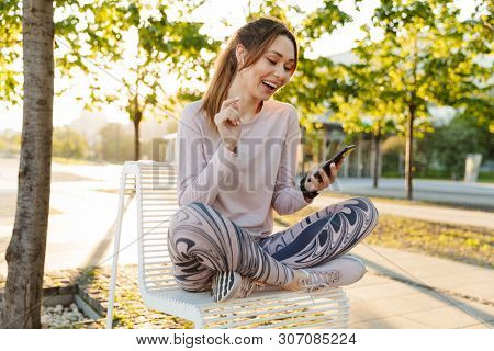 Cheerful young fitness woman relaxing outdoos, sitting on a bench, using mobile phone