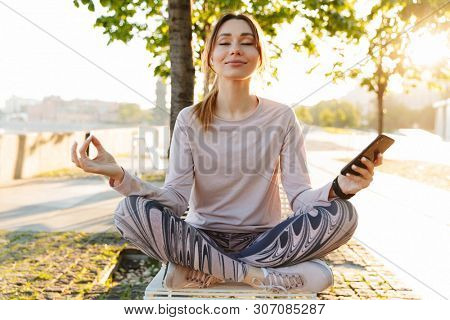 Cheerful young fitness woman relaxing outdoos, sitting on a bench, using mobile phone, meditating