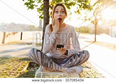 Cheerful excited young fitness woman relaxing outdoos, sitting on a bench, using mobile phone