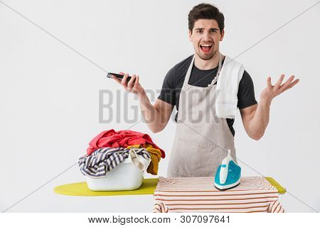 Image of a young confused displeased houseman househusband with iron near clothes isolated over white wall background using mobile phone.