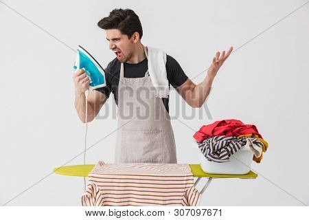 Image of a young angry houseman househusband screaming on iron near clothes isolated over white wall background.