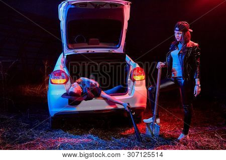 Female Bandit Near Car With A Shovel And A Dead Or Drunk Guy In The Trunk At Night. Photoshoot About
