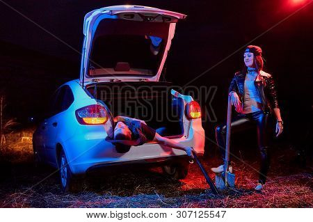 Female Bandit Near Car With A Shovel And A Dead Or Drunk Guy In The Trunk At Night. Photoshoot About