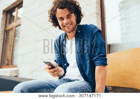 Smiling Young Man Stitting Outdoors Texting On Mobile Phone. Happy Male With Curly Hair Using Smart 