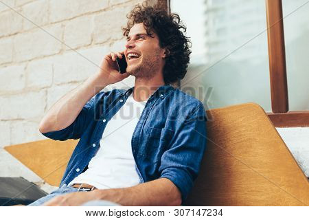 Young Caucasian Man Stitting On The Bench Outdoors And Talking On Mobile Phone. Happy Male With Curl