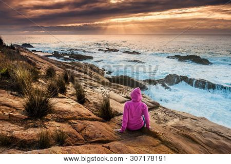 Female In Pink Hoodie Relaxes On Coastal Rocks Watching The Sunbeams And Sea Flows Over The Craggy R