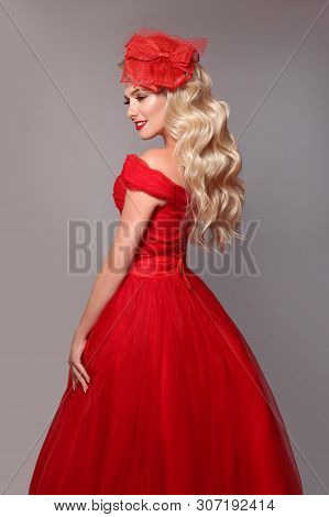 Blonde Woman In Red Dress And Elegant Hat Isolated On Studio Grey Background. Beautiful Blond Girk W