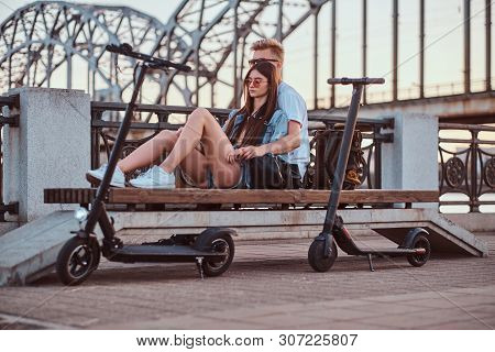 Young Beautiful Romantic Couple With Electric Scooters Is Sitting On The Bench Near Brige At Sunset 