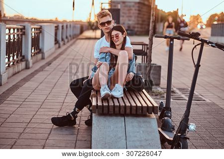 Young Beautiful Romantic Couple With Electric Scooters Is Sitting On The Bench Near Brige At Sunset 