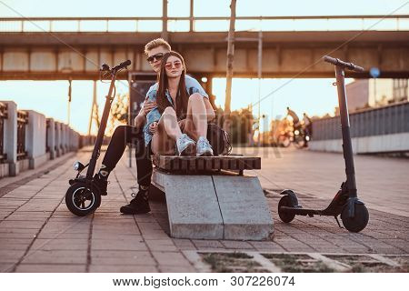 Young Beautiful Romantic Couple With Electric Scooters Is Sitting On The Bench Near Brige At Sunset 