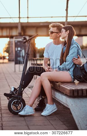 Young Beautiful Romantic Couple With Electric Scooters Is Sitting On The Bench Near Brige At Sunset 