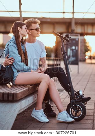 Young Beautiful Romantic Couple With Electric Scooters Is Sitting On The Bench Near Brige At Sunset 