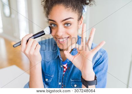 Young african american girl appying blush using make up brush doing ok sign with fingers, excellent symbol