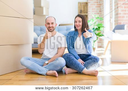 Young couple sitting on the floor arround cardboard boxes moving to a new house doing happy thumbs up gesture with hand. Approving expression looking at the camera with showing success.