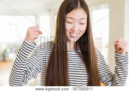 Beautiful Asian woman wearing stripes sweater over living room background excited for success with arms raised celebrating victory smiling. Winner concept.