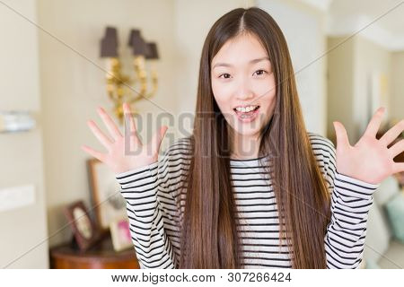 Beautiful Asian woman wearing stripes sweater over living room background celebrating crazy and amazed for success with arms raised and open eyes screaming excited. Winner concept