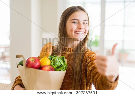 Beautiful young girl holding paper bag of fresh groceries happy with big smile doing ok sign, thumb up with fingers, excellent sign