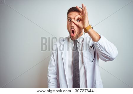 Young handsome doctor man wearing white profressional coat over isolated background doing ok gesture shocked with surprised face, eye looking through fingers. Unbelieving expression.