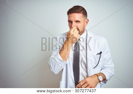 Young handsome doctor man wearing white profressional coat over isolated background smelling something stinky and disgusting, intolerable smell, holding breath with fingers on nose. Bad smells concept