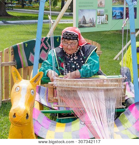 Yekaterinburg, Russia, June 15, 2019. An Elderly Woman In Bashkir Clothes Is Seated At A Old Wooden 