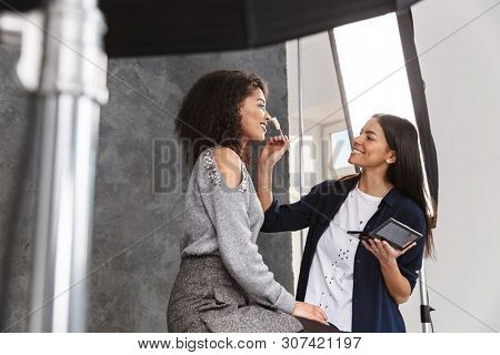 Portrait of female makeup artist applying cosmetics to beautiful woman during photo shooting with professional camera and softbox in studio