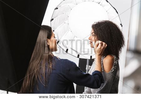 Portrait of female makeup artist applying cosmetics to gorgeous woman during photo shooting with professional camera and softbox in studio
