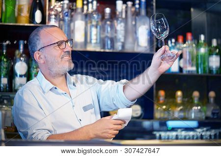 Elderly Bartender Cleans The Glass. A Handsome Bartender Polishes A Glass Of Wine Glasses. The Conce
