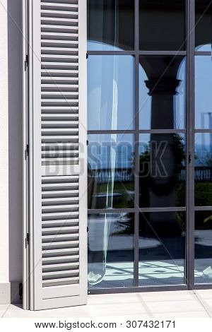 Wooden Shutters Of Gray Wood Are Open To Sunlight Near The Panoramic Windows With A Black Frame, Clo
