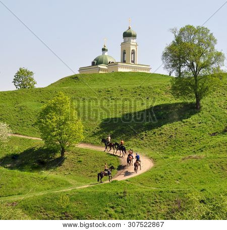 Green Hilly Terrain In Spring. Horsemen Are Riding Along A Country Road.